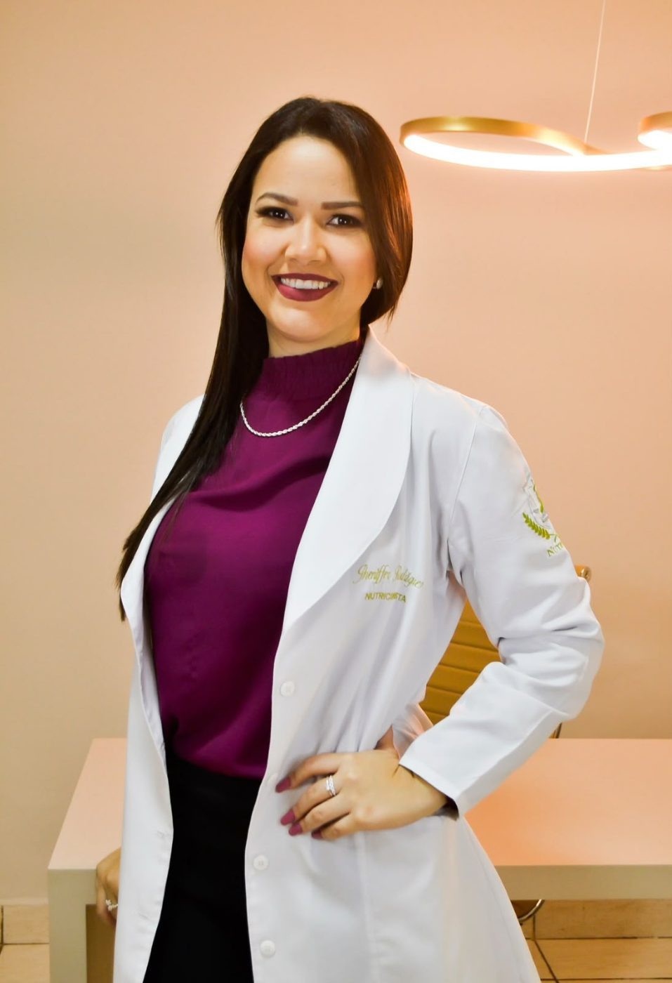 A nutricionista Jheniffer Rodrigues, de jaleco, sorrindo no consultório